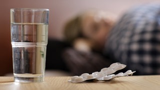 Eine junge Frau liegt krank im Bett, auf dem Nachttisch steht ein Glas Wasser und daneben Schmerztabletten.