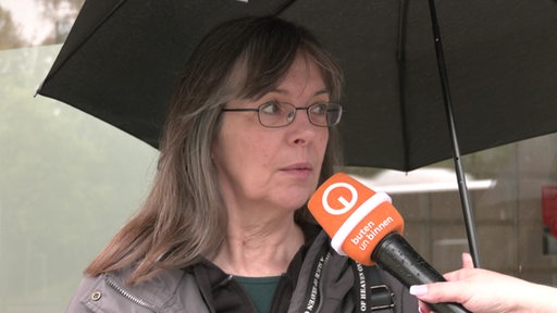 Eine Frau steht unter einem schwarzen Regenschirm und wird interviewt.