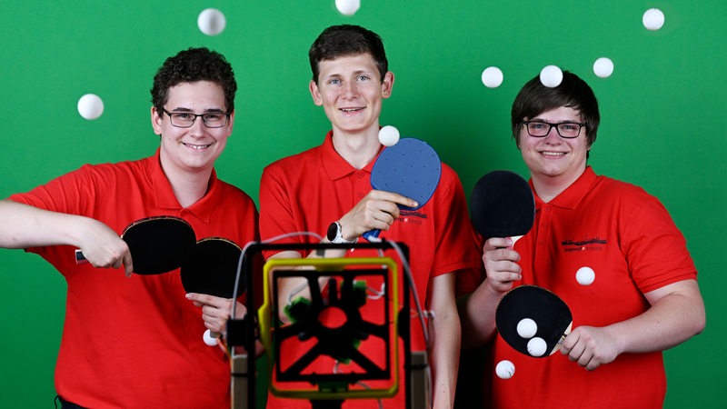 Drei Schüler, die bei "Jugend forscht" gewonnen haben, stehen inmitten von Tischtennisbällen.