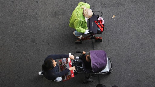 Eine alte Frau mit Rollator geht neben einer Mutter, die einen Kinderbuggy schiebt über die Straße. Als Aufsicht fotografiert