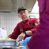 Ein Mann mit roter Schürze und Polizemütze steht in einer Küche.