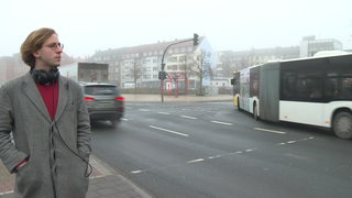 Karl Geibert steht mit Kopfhörern an einer stark befahrenen Hauptstraße.