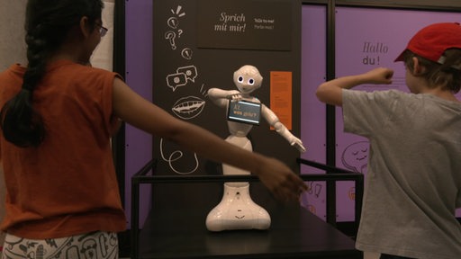 Zwei Kinder stehen vor einem kleinen Roboter, welcher ihre Bewegungen nachahmt.