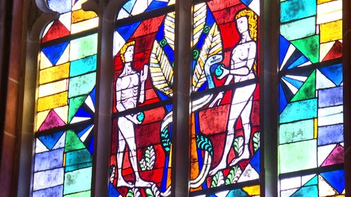 Ein buntes Kirchenfenster auf dem eine Szene aus der Bibel mit Adam und Eva zu sehen ist. 