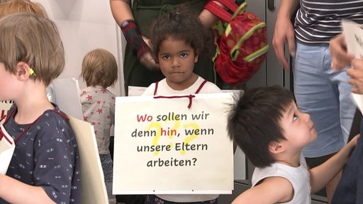 Ein Kind steht mit einem Schild der Aufschrift "Wo sollen wir denn hin, wenn unsere Eltern arbeiten" in einer Kita.