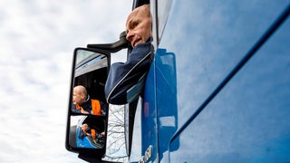 Ein Lkw-Fahrer rangiert seinen Laster rückwärts. Dabei schaut er aus dem Fenstern nach hinten und spiegelt sich in seinem Seitenspiegel.