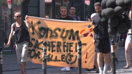 Zwei Passanten halten ein Plakat mit der Aufschrift "Konsum Sicherheit für alle".