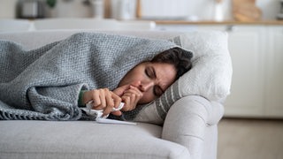 Eine kranke Frau liegt auf der Couch, eingewickelt in eine Decke, und hustet.