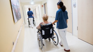2 Pflegerinnen schieben älteren Patientinnen im Rollstuhl über einen Krankenhausflur.