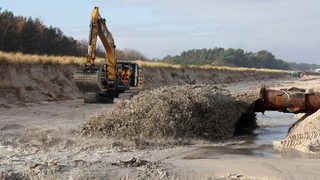 Vor einem beschädigten Deich schießt ein Wasser-Sand-Gemisch aus einem Rohr, dahinter steht ein Bagger bereit. 