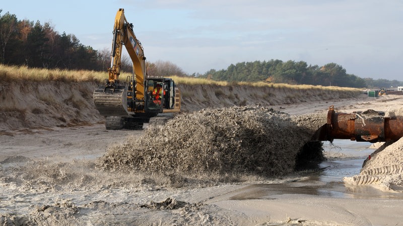 Vor einem beschädigten Deich schießt ein Wasser-Sand-Gemisch aus einem Rohr, dahinter steht ein Bagger bereit. 