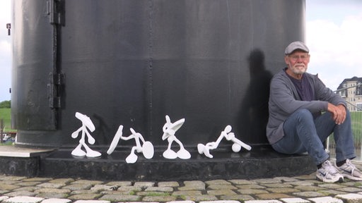 Der Künstler und Holzbildhauer Rainer Madena sitzt neben vier kleinen weißen Skulpturen.