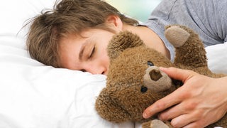 Ein junger Mann liegt schlafend mit seinem Teddy im Arm im Bett.