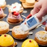 Ein Lebensmittelkontrolleur hält in einer Patisserie bei einer Kontrolle ein Thermometer an eine kleine Torte. 