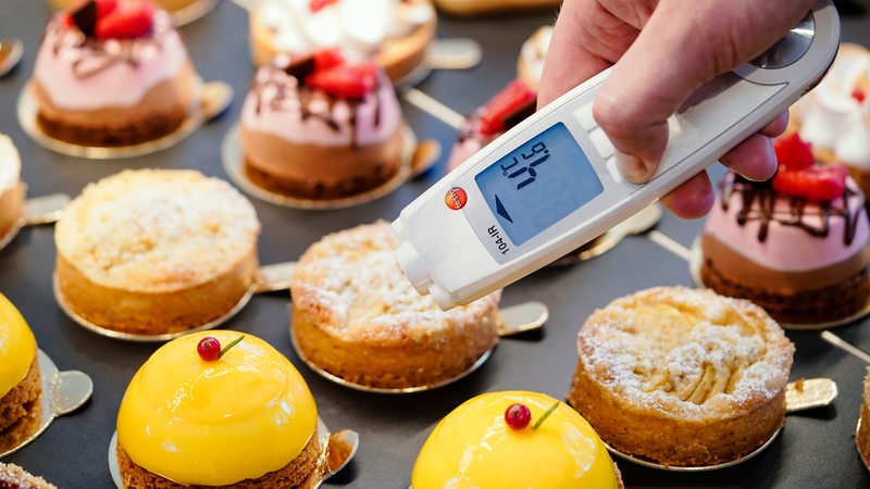 Ein Lebensmittelkontrolleur hält in einer Patisserie bei einer Kontrolle ein Thermometer an eine kleine Torte. 