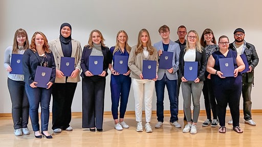 Zwölf Menschen stehen nebeneinander auf einer Bühne und halten jeweils eine blaue Mappe in der Hand.