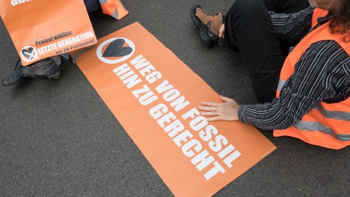Ein Protestschild mit der Aufschrift "Weg von Fossil hin zu gerecht" liegt auf dem Boden, daneben sitzt ein Mann