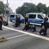 Sieben Menschen sitzen auf einer Straße, Polizisten stehen daneben und dahinter, ebenso Polizeiwagen.