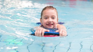 Ein Mädchen im Schwimmbad klammert sich an ein Schwimmbrett und schwimmt in Richtung Kamera. 