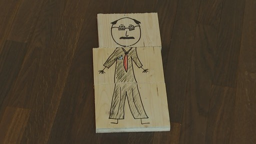 Auf Holzstücken ist ein Mann mit Brille und Anzug gezeichnet.