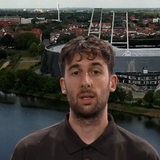 Ein junger Mann schaut in eine Kamera. Hinter ihm ein Stadion.