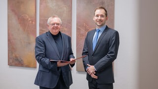 Zwei Männer im Anzug stehen nebeneinander, einer hält eine Mappe in der Hand.