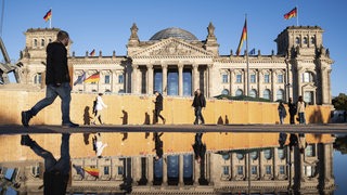 Menschen gehen an einem sonnigen Herbsttag am Reichstag im Berliner Bezirk Mitte vorbei, dessen Westfassade sich in einer großen Pfütze davor spiegelt. 