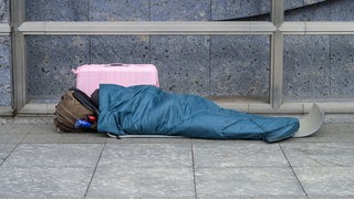 Ein obdachloser Mann liegt in einem Schlafsack, daneben steht einer Koffer