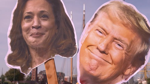 Zwei ausgeschnittene Köpfe aus Pappe, befestigt auf Holzstilen. Der Linke Kopf ist Kamala Harris und der rechte Donald Trump.