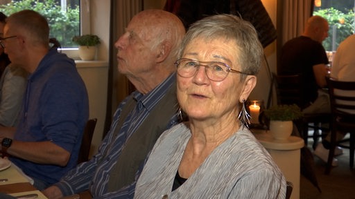 Eine ältere Frau mit kurzen grauen Haaren und Brille schaut in die Kamera.