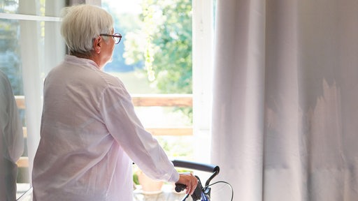 Alte Frau als Patientin mit Rollator in der Reha Klinik schaut aus dem Fenster in den Sonnenschein