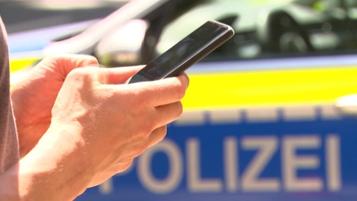 In zwei Händen wird ein Smartphone gehalten und im Hintergund lässte sich ein Polizeiwagen erkennen.