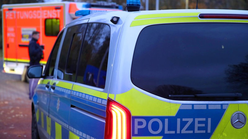 Ein Polizeiauto fährt mit Blaulicht durch eine Straße zum Einsatz. Im Hintergrund ist ein Fahrzeug des Rettungsdienstes zu sehen.