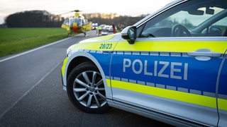 Ein Polizeiwagen steht auf einer Landstraße. Im Hintergrund ist ein Rettungshubschrauber zu sehen.