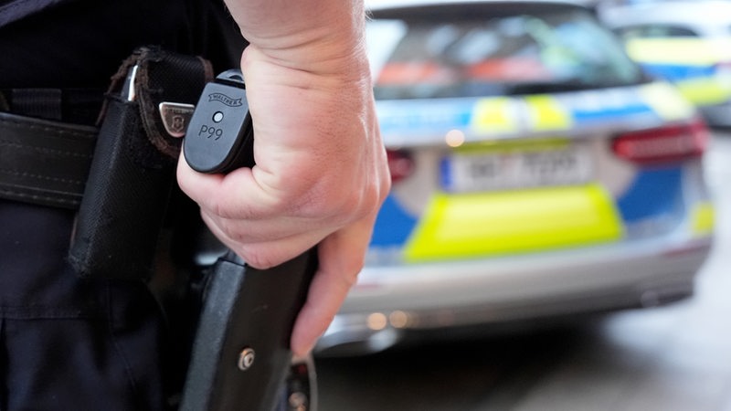 Ein Polizist steht mit seiner Hand auf der Schusswaffe vor einem Einsatzfahrzeug der Polizei.