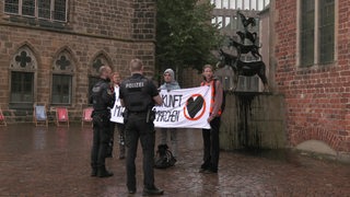 Zwei Polizisten stehen vor Aktivisten, die vor den mit Farbe übergossenen Bremer Stadtmusikanten ein Banner hochhalten.