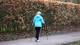 Eine Frau läuft auf einem Weg neben einer Hecke mit braunen Bättern. Sie trägt eine hellblaue Jacke, eine schwarze Hose und läuft mit zwei Nordic-Walking-Stöcken-