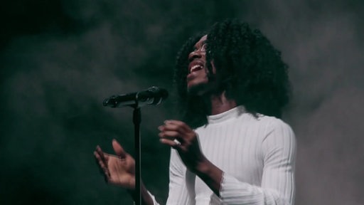 Ein Sänger mit dunklen Locken und weißem Top steht auf einer Bühne vor einem Mikrofon