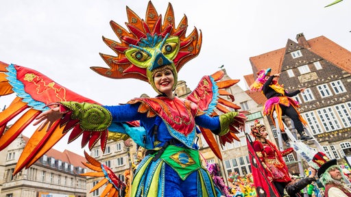 Stelzenläuferin mit bunter Maske und Flügel-Kostüm tanzt auf dem Bremer Marktplatz.