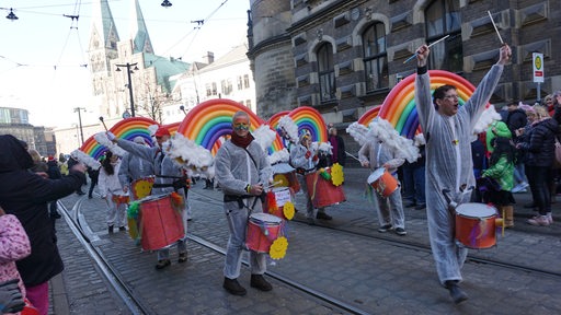 Eine Trommeltruppe in weißen Overalls. Auf ihrem Rücken sind Schwimmnudeln in unterscheidlichen Farben befestigt. Sie sollen an einen Regenbogen erinnern.