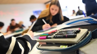 Stifte liegen in einem Etui in einem Klassenzimmer, Schülerinnen schreiben im Hintergrund.