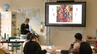 In einem Klassenraum in einer Schule sitzen mehrere Schüler an Tischen, während eine Lehrerin vor einer Leinwand steht.