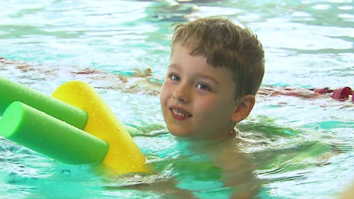 Ein Kind hält sich während eines Schwimmkurses an einer Schwimmnudel fest und lächelt in die Kamera