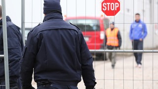 Mitarbeiter eines Sicherheitsdienstes sind von hinten zu sehen, wie sie einen Bauzuan passieren.