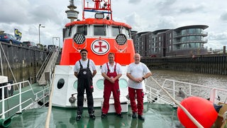 Drei Männer stehen an Deck eines Seenotrettungskreuzers im Hafen.