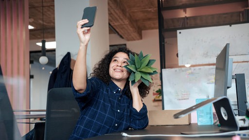 Eine Frau sitzt vor einem Laptop und macht ein Selfie, während sie eine Pflanze in der Hand hält.