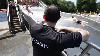 Ein Mann von einem Sicherheitsdienst steht im Freibad an einer Wasserrutsche.