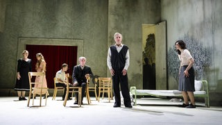 Foto von einer Szene im Theater mit sechs Männern und Frauen im Vordergrund einer angedeuteten Wohnung