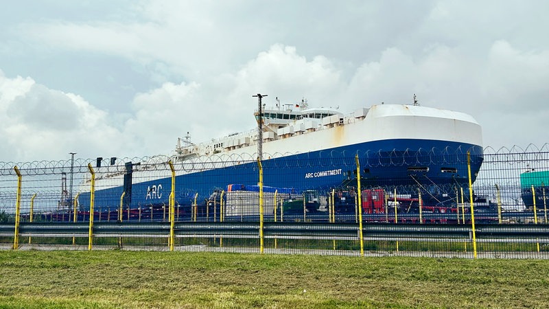 Hinter einem Zaun liegt ein großes blaues Frachtschiff.