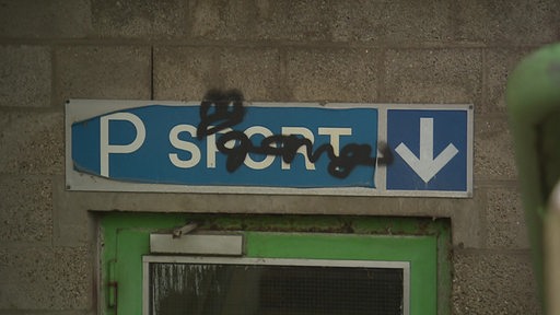 Das marode Eingangsschild einer Sporthalle, auf dem ein Grafitti prangt. 